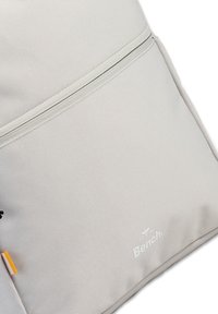 Hellgrauer Rucksack mit einer strukturierten Oberfläche, ausgestattet mit einer Reißverschlusstasche, einem dezenten Logo und einem orangefarbenen Etikett.