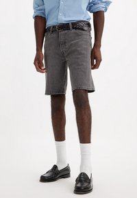 Hombre con camisa de manga larga azul claro arremangada, cinturón de cuero negro trenzado, pantalones cortos de mezclilla negra deslavada, calcetines blancos hasta la altura del tobillo y zapatos mocasines de cuero negro.