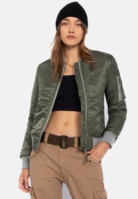 Olivgrüne Bomberjacke mit satinierter Oberfläche, gerippten Bündchen, Reißverschlusstaschen und einem Frontreißverschluss. Getragen über einem schwarzen Crop-Top und beigen Cargo-Hosen.