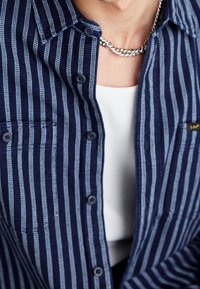 Närbild av en person som bär en blå och vit randig skjorta med knappar över en vit linne med ett silverfärgat kedjehalsband.