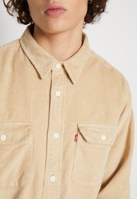 Lichtbruine corduroy blouse met een kraag, voorzien van twee borstzakken, witte knopen en een klein rood Levi's logo label aan de zoom.