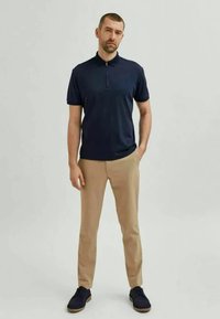 Jūras spēku polo krekls ar rāvējslēdzēju, apvienots ar bēšiem biksēm un tumšām kurpēm. Apģērbs ir ar pieguļošu dizainu un gludu tekstūru.