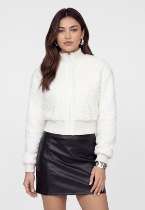 Femme portant une veste blanche texturée zippée et une jupe en cuir noire, debout devant un fond blanc.