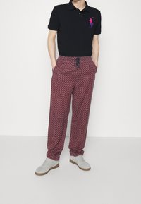 Svart polo-shirt med en liten flerfärgad logga, kombinerad med vinröda mönstrade svettbyxor och grå sneakers med vita accenter.