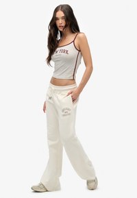 Joven mujer vestida con un crop top gris de Nueva York, pantalones de chándal crema de Superdry Athletics y zapatillas beige, de pie con una mano en el bolsillo.