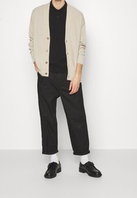 Cardigan beige avec une texture côtelée, polo noir, pantalon noir à coupe décontractée, chaussures noires avec un détail boucle et chaussettes blanches.