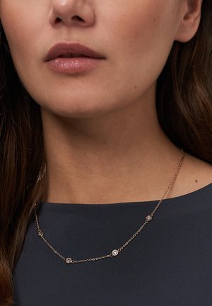 Nærbillede af en kvindes nedre del af ansigtet og nakke, der bærer en guldkæde med fire små runde klare ædelstene over en mørk top.