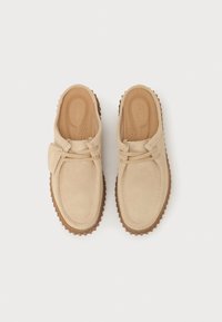 Beige suede shoes with a slip-on design, featuring two laces across the front, cushioned insole, and a textured brown rubber sole.