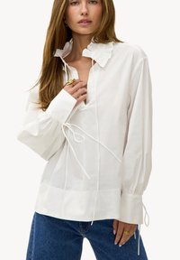 Femme portant un chemisier blanc ample avec un col à volants et des manches longues bouffantes, associé à un jean bleu et des bagues en or.