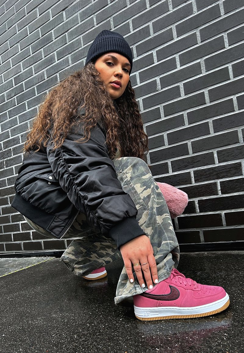 Jeune femme aux longs cheveux bouclés, portant un bonnet noir, une veste noire, un pantalon camouflage et des baskets roses, posant accroupie contre un mur de briques sombre.