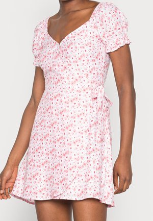 Robe fluide blanche à fleurs roses, avec des manches courtes bouffantes, un décolleté en V et une taille nouée. Tissu léger avec une jupe ample.