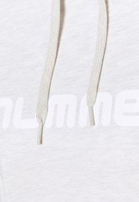 Gros plan sur un tissu de hoodie blanc avec des cordons de serrage blancs et un lettrage en gras blanc partiellement visible.