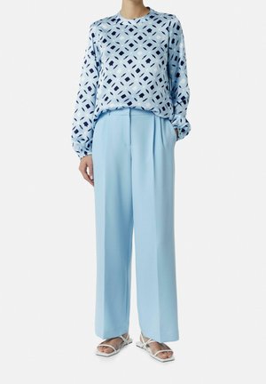 Mujer que lleva pantalones anchos azul claro, una blusa azul de manga larga con estampado y sandalias blancas abiertas de dedo, de pie con las manos en los bolsillos.