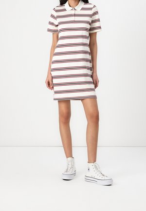 Kobieta ubrana w krótką sukienkę polo z kołnierzykiem i paskami, nosząca białe wysokie trampki, na białym tle.