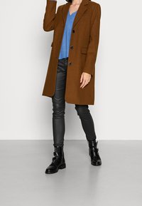 Brauner Wollmantel mit Reverskragen und fünf schwarzen Knöpfen, kombiniert mit einem blauen Strickpullover und schwarzen Lederhosen, getragen mit schwarzen Stiefeletten.
