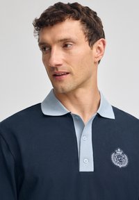 Mörkblå polo skjorta med ljusblå krage, två-knapplock, och broderad emblem på vänster bröst. Slät bomullstyg.