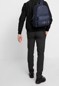 Mies, joka on pukeutunut mustaan takkiin, harmaisiin housuihin ja mustiin kenkiin, kantaa tummansinisessä Lacoste-repussa, jossa on värikkäät logokirjaimet, olallaan.