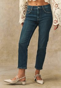 Jeans de denim azul oscuro con corte recto y dobladillo sin acabar, combinados con zapatos de salón en color crema con punta afilada y finas correas en el tobillo.