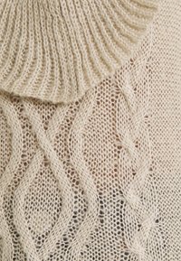 Pull en maille beige avec un col côtelé et des motifs torsadés, présentant une texture douce avec des mailles ouvertes et des variations subtiles.