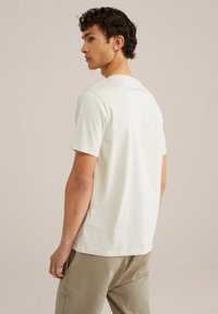 Tricou crem cu mânecă scurtă și decolteu rotund, realizat dintr-un material moale; asociat cu pantaloni gri deschis, văzut din spate.