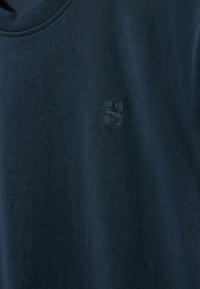 Tums zilas kokvilnas T-krekls ar apaļu izgriezumu un teksturētu izšūtu "S" logo kreisajā krūšu daļā. Mīksta, gluda tekstūra.