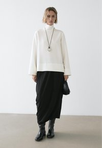 Weißes langärmliges Rollkragenshirt kombiniert mit einem schwarzen, drapierten Rock. Das Outfit wird durch eine schwarze Tasche und kniehohe schwarze Stiefel ergänzt.