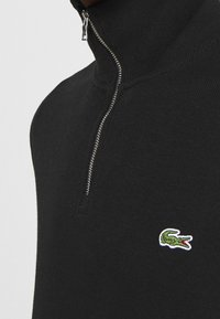 Maglione nero con collo a zip in tessuto a coste, con piccolo logo coccodrillo verde ricamato sul petto. Dettaglio della cerniera in metallo color argento.
