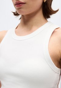 Κοντινό πλάνο μιας γυναίκας που φοράει λευκό αμάνικο πλεκτό τοπ και χρυσούς κρίκους στα αυτιά, εμφανίζοντας τον λαιμό, τα χείλη και τον ώμο της.