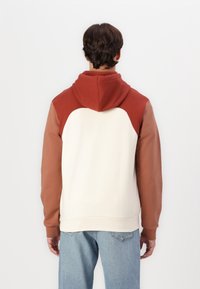 Sudadera con capucha en color crema, con acentos en óxido y marrón, que presenta un corte holgado y puños acanalados. Textura suave, bloques de color sólido.