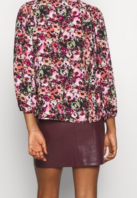 La blouse florale présente un motif vibrant en rose, vert et noir avec des manches bouffantes 3/4. Associée à une jupe en cuir bourgogne courte.