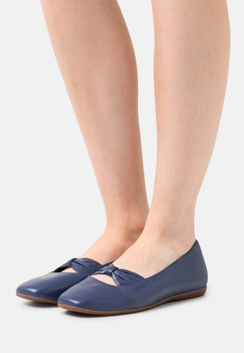 Pieds portant des ballerines en cuir bleu marine avec un détail de nœud torsadé et des semelles basses marron, photographiés sur un fond clair.