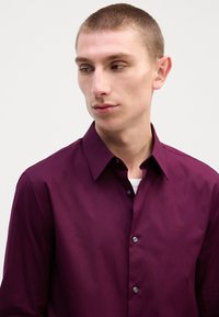 Mørk lilla skjorte med knapp ned, laget av glatt stoff, med klassisk krage og svarte knapper, samt en liten logo på venstre side.