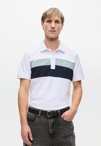 Valkoinen polo paita, jossa on tummansininen ja vaaleanvihreä vaakaraita, lyhyet hihat ja klassinen kaulus, yhdistettynä tummiin farkkuihin.