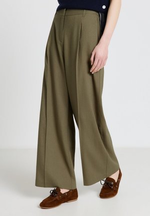 Persona che indossa pantaloni larghi verde oliva e scarpe marroni in camoscio con lacci, in piedi su un pavimento bianco.