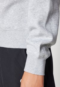 Felpa grigia chiara con tessuto testurizzato, polsini a costine e scollatura rotonda. Indossata con pantaloni scuri, che mettono in evidenza il dettaglio delle maniche.