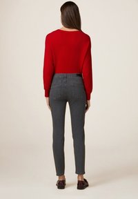 Femme se tenant de dos, portant un pull rouge, un jean gris ajusté et des mocassins marron foncé, sur un fond uni.