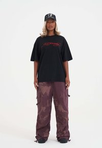 Sort oversized t-shirt med røde broderede bogstaver parret med maroon cargo bukser med et mønster af hjerter og drager. En sort cap fuldender looket.