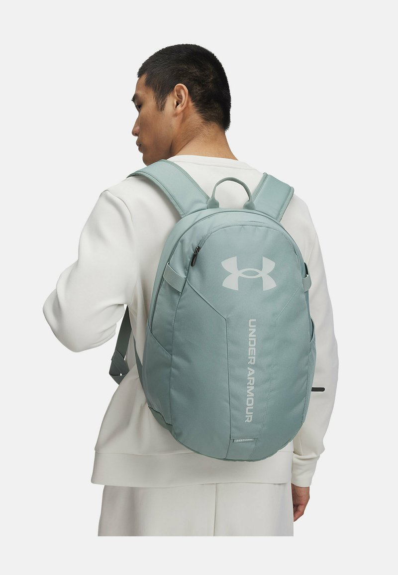 Zaino Under Armour azzurro chiaro con una texture liscia, spallacci regolabili, maniglia superiore e tasca con zip laterale. Presenta il logo sul lato frontale.