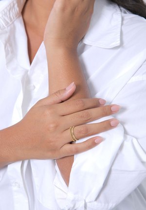 Anello a doppia fascia in oro con un design texture, indossato su una mano con pelle chiara e unghie curate di un rosa chiaro, su una camicia bianca.