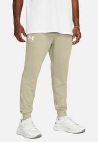 Pantalones de chándal beige con puños acanalados, con un logo de Under Armour en blanco en el muslo izquierdo. Llevados con zapatillas deportivas blancas.