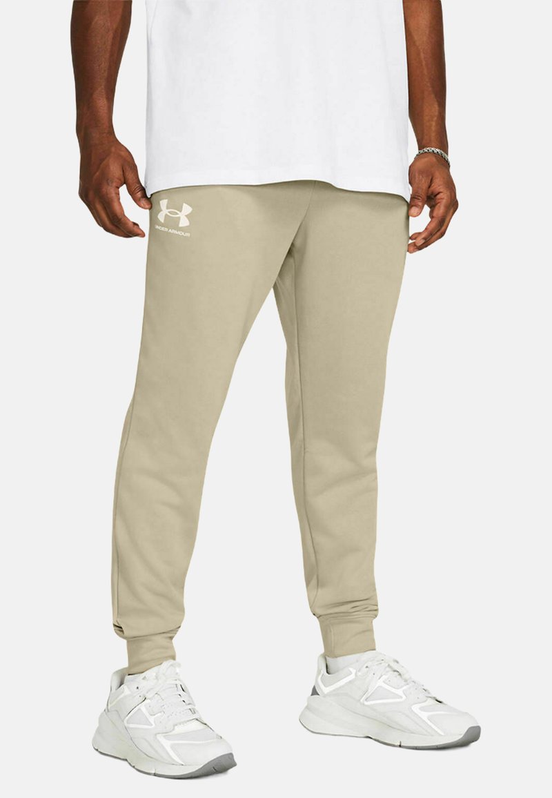 Pantalones de chándal beige con puños acanalados, con un logo de Under Armour en blanco en el muslo izquierdo. Llevados con zapatillas deportivas blancas.