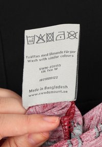 Tvättetikett på lätt tyg, med tvättanvisningar och ikoner. Vit med svart text. Taggen innehåller varumärkes- och ursprungsinformation.