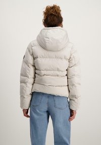 Een pufferjack in lichtbeige met een capuchon, met gestikte secties en een boxy vorm. Draagt over blauwe jeans, gezien van achteren.