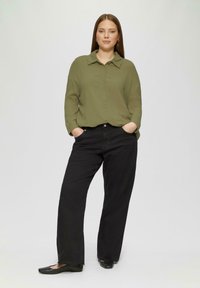 Olijfgroene overhemd met knopen en een kraag, zwarte wijde broek en platte zwarte schoenen. Model staat met handen in de zakken op een lichte achtergrond.