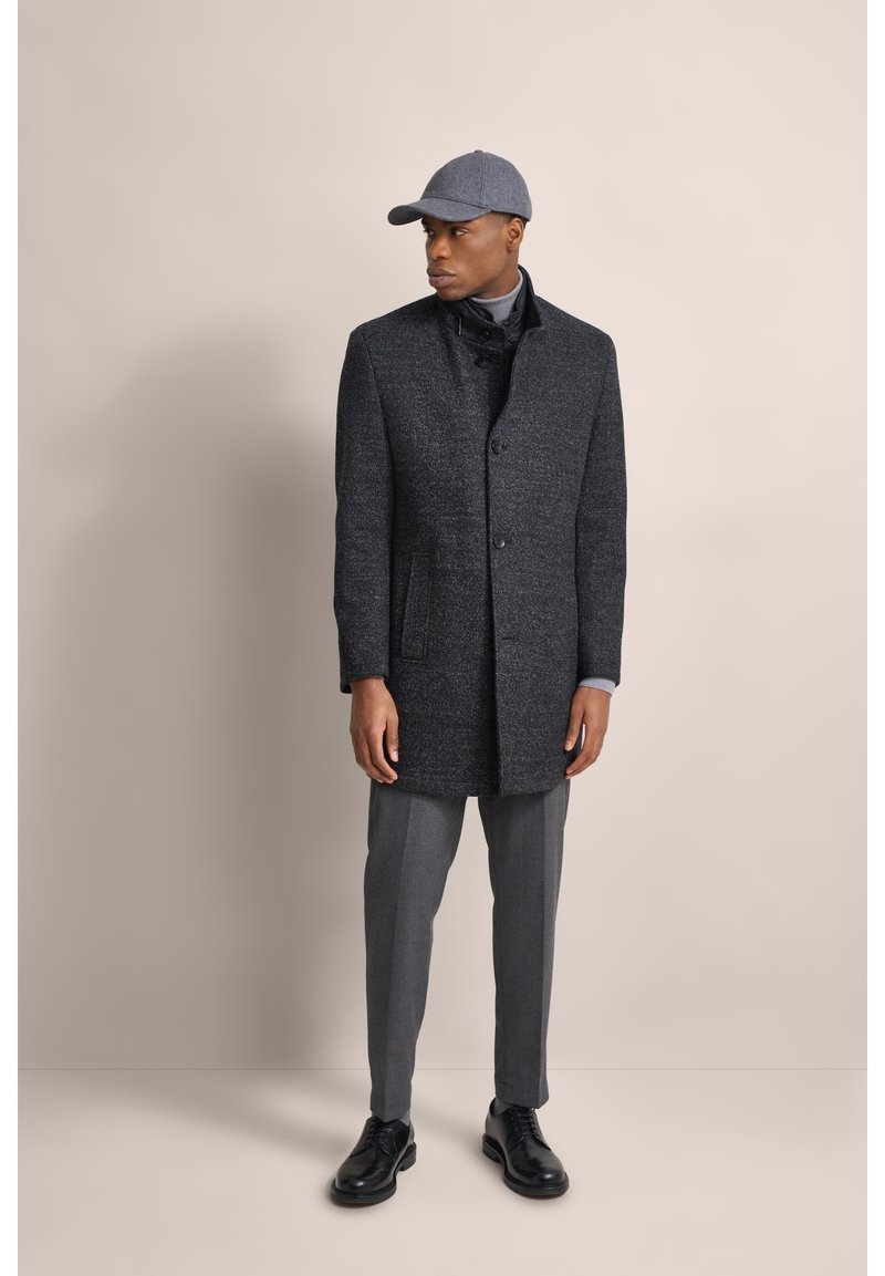 Cappotto di lana grigio scuro con una finitura testurizzata e chiusura con bottone, abbinato a pantaloni grigi e scarpe nere, indossato con un cappellino grigio.