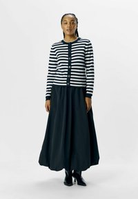 Cardigan rayé en bleu marine et blanc, avec un col rond et une fermeture à boutons, associé à une longue jupe noire et des bottines noires.