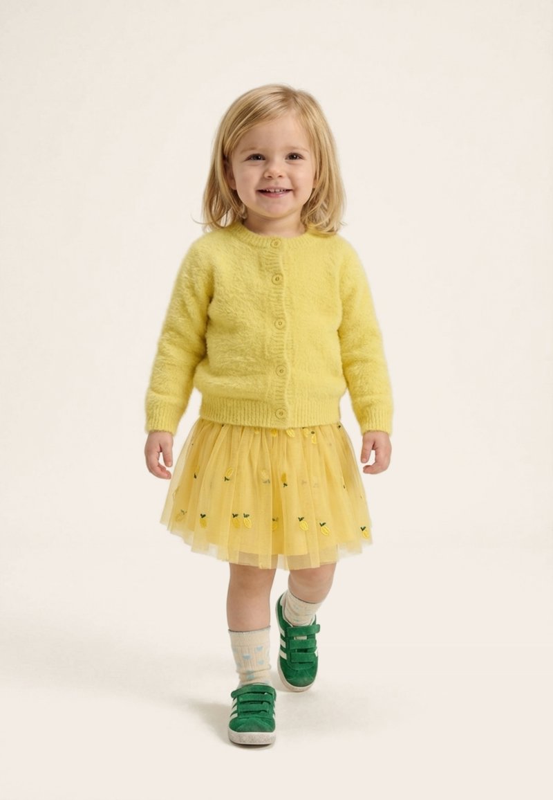 Jeune fille souriante marchant, portant un cardigan jaune duveteux, une jupe en tulle jaune brodée, des chaussettes dépareillées et des baskets vertes, sur un fond uni.