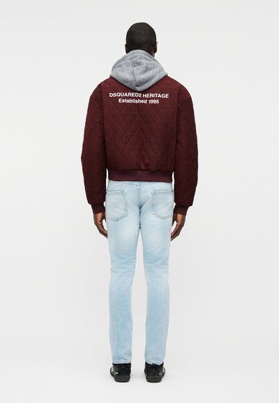 Homme portant une veste matelassée bordeaux avec le texte blanc « DSQUARED2 HERITAGE Established 1995 », un sweat à capuche gris, un jean bleu clair et des chaussures noires, vu de dos.