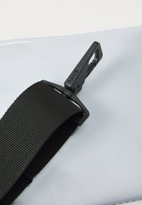 Bretelle noire réglable avec une boucle en plastique, matériau texturé, fixée à une surface lisse et brillante de couleur gris clair. "RANS" embossé sur la boucle.