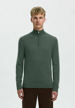 Green knit sweater with a high collar and half zip. Features long sleeves and a smooth texture. Paired with brown corduroy pants.